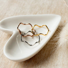 Load image into Gallery viewer, Three wavy metal rings in rose gold, yellow gold, and silver tones arranged on a small white ceramic dish over a light wood surface with soft decorative fibers nearby.