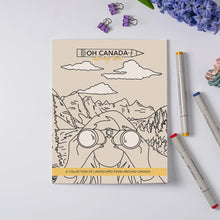 Load image into Gallery viewer, A flat-lay of a coloring book titled 'OH CANADA - COLORING PAGE COLLECTION FROM AROUND CANADA' on a light gray background, styled with purple flowers, binder clips, and three markers.
