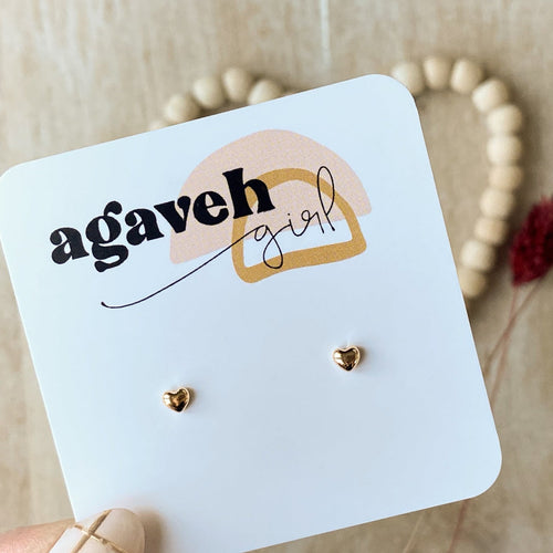 A pair of tiny gold heart-shaped stud earrings displayed on a white card with 'agaveh girl' branding, accompanied by a beaded necklace and decorative elements in the background.